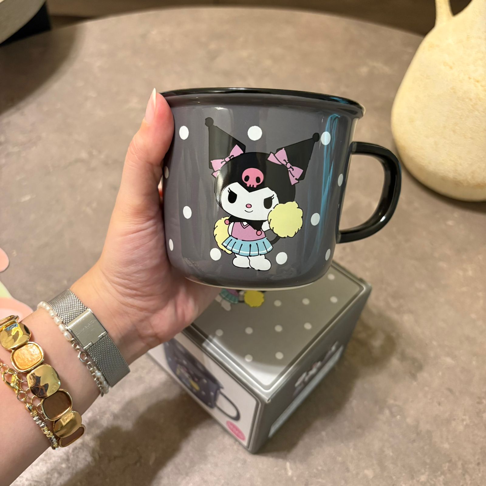 A hand holds a dark gray enamel mug featuring a cartoon character with a pink bow and black polka-dot hat. The mug has a black handle and rim, and the background shows a gray stone surface with a matching box underneath.
