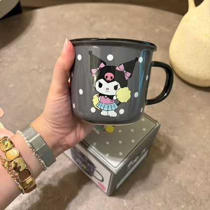 A hand holds a dark gray enamel mug featuring a cartoon character with a pink bow and black polka-dot hat. The mug has a black handle and rim, and the background shows a gray stone surface with a matching box underneath.