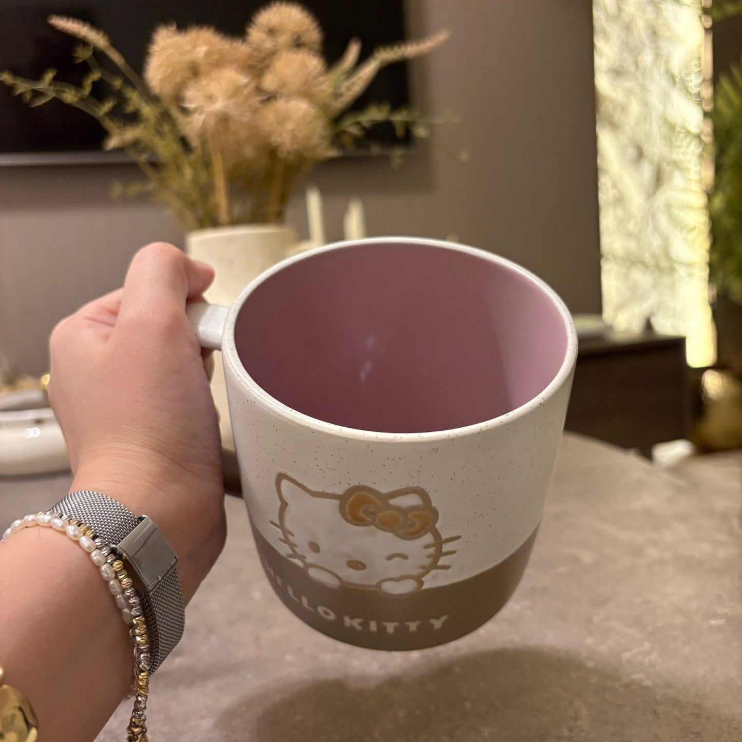 A hand holding a Hello Kitty ceramic mug with a speckled off-white exterior, a dusty pink interior, and a brown bottom section featuring the Hello Kitty face and 'HELLO KITTY' text, set against a blurred indoor background.