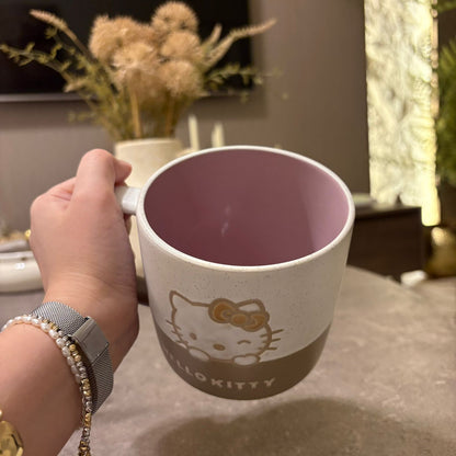A hand holding a Hello Kitty ceramic mug with a speckled off-white exterior, a dusty pink interior, and a brown bottom section featuring the Hello Kitty face and 'HELLO KITTY' text, set against a blurred indoor background.