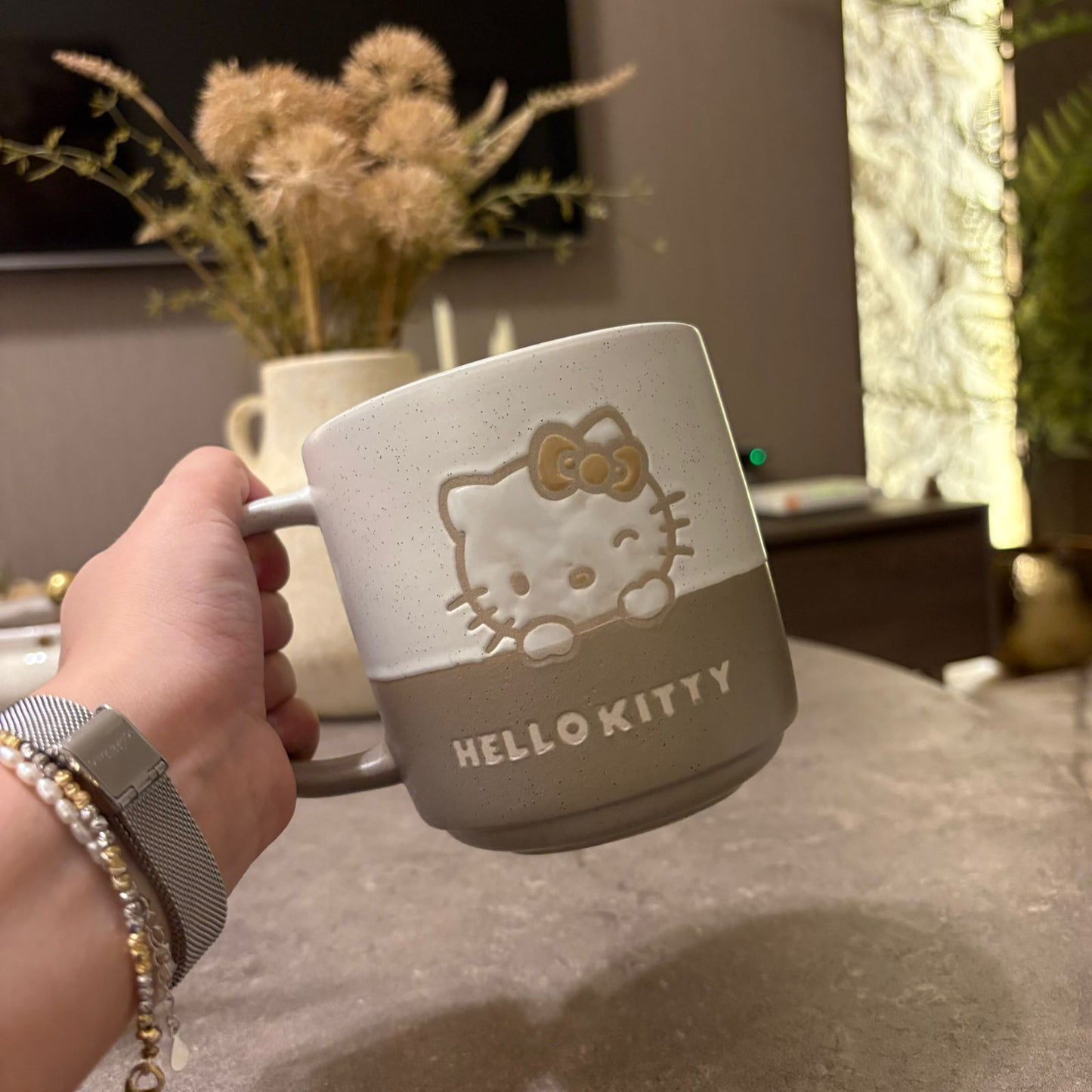 A hand holds a speckled ceramic Hello Kitty mug with a two-tone beige and gray design and a raised Hello Kitty face. The background shows a cozy indoor setting with a blurred vase of dried flowers and a table.