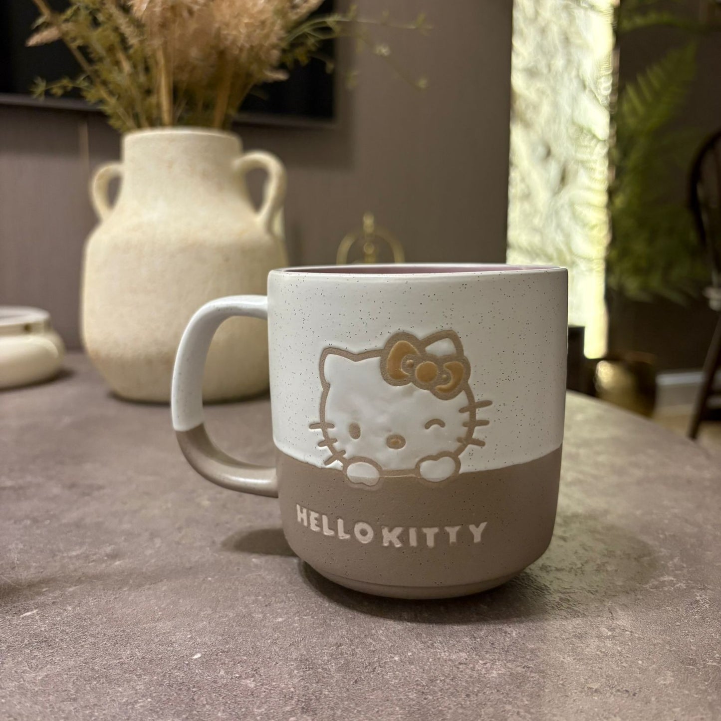 A ceramic Hello Kitty mug with a speckled off-white upper half and a taupe-brown lower half, featuring an embossed Hello Kitty face and bow on the front, set on a gray tabletop with a blurred vase in the background.
