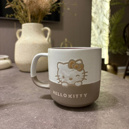 A ceramic Hello Kitty mug with a speckled off-white upper half and a taupe-brown lower half, featuring an embossed Hello Kitty face and bow on the front, set on a gray tabletop with a blurred vase in the background.