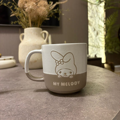 A ceramic mug with a two-tone beige and off-white design, featuring an embossed bunny character and the text 'MY MELOODY' on the front, sitting on a gray stone tabletop with a blurred background.