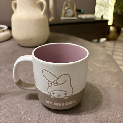 A ceramic mug with a white exterior, a light brown lower half featuring a My Melody cartoon design, and a dusty purple interior, set on a gray tabletop.