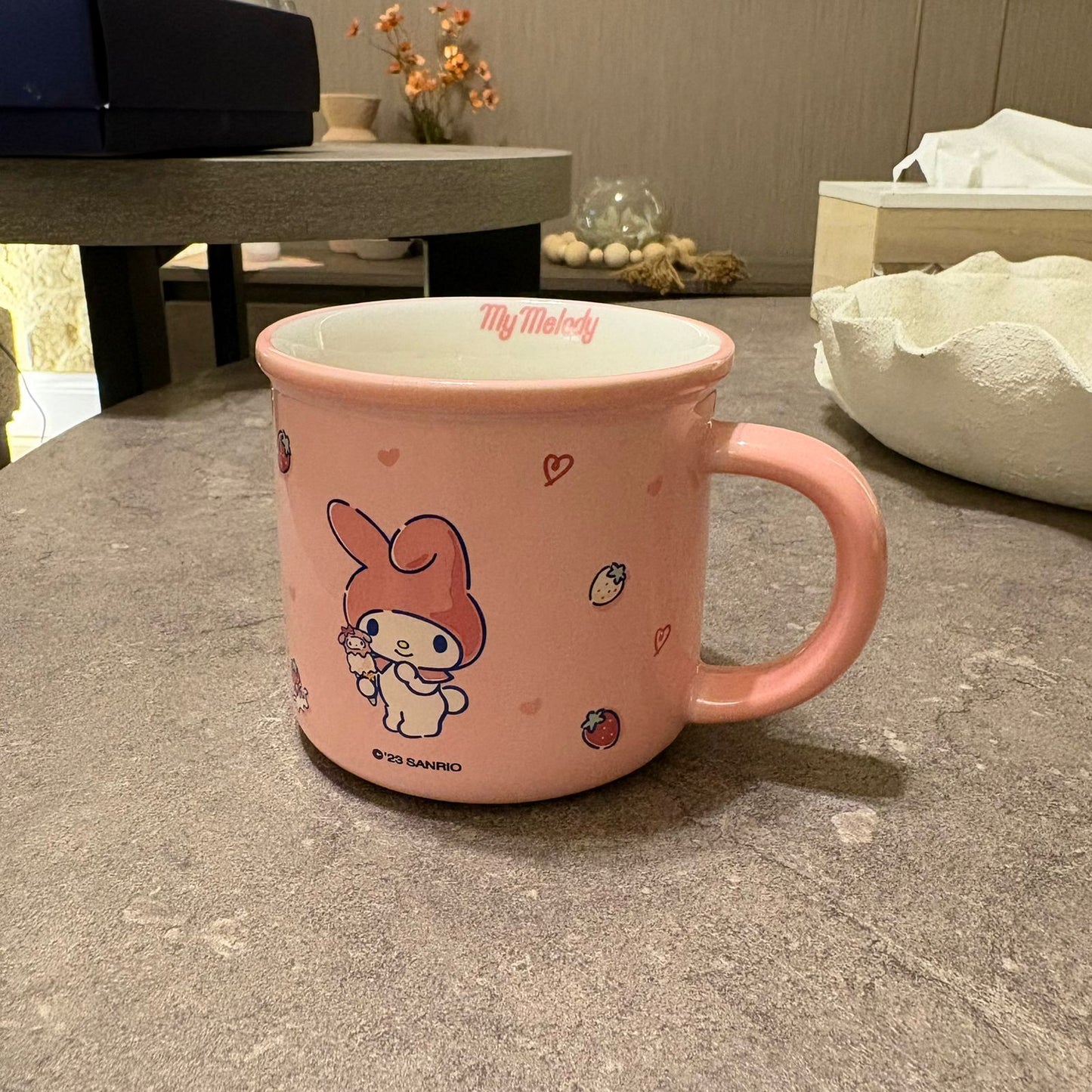 A pink ceramic mug featuring a My Melody character illustration and the text 'My Melody' and '© PD SANRIO' on a gray tabletop in a home setting.