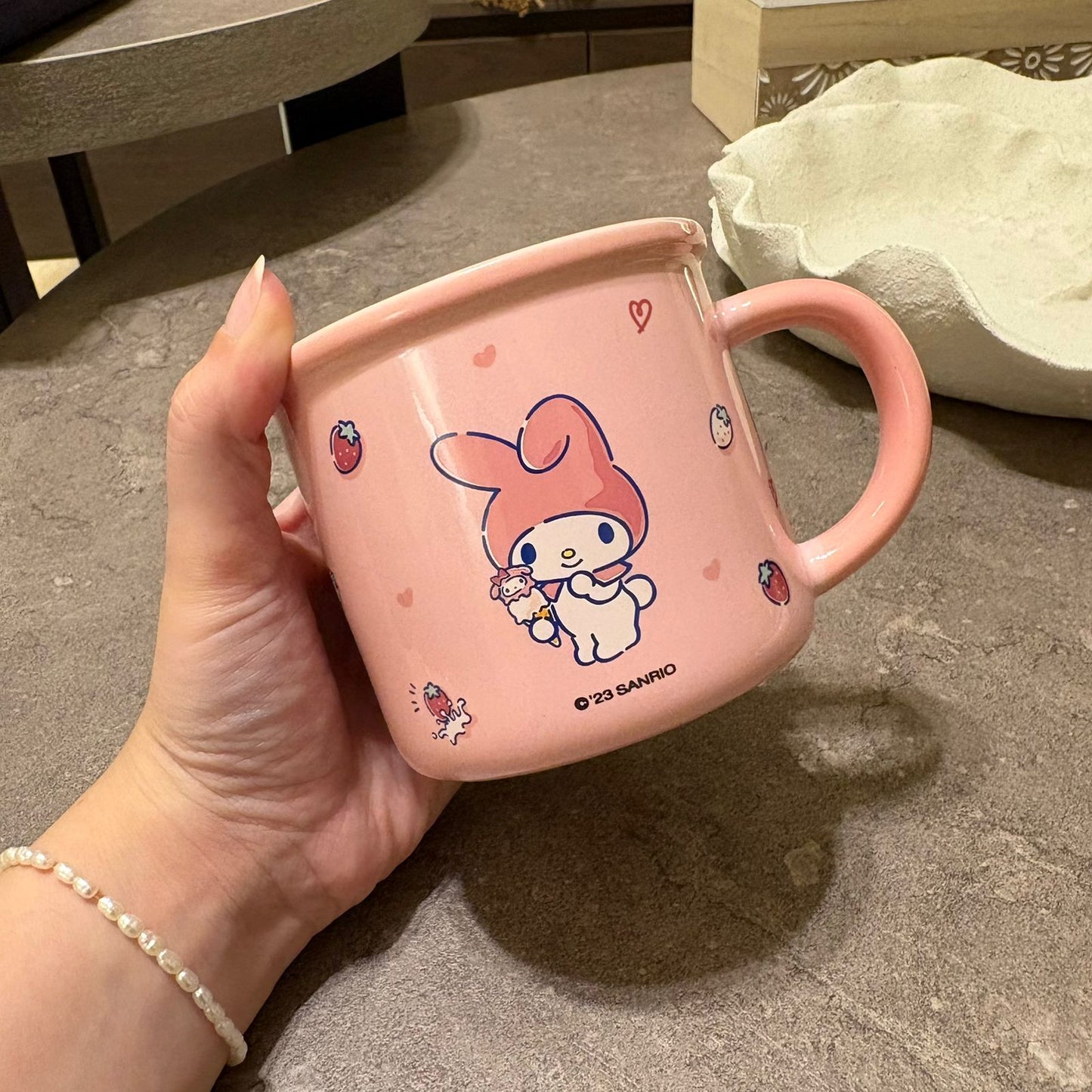 A hand holds a pink ceramic mug featuring the Sanrio character My Melody, with strawberry and heart details, set against a gray stone tabletop background.