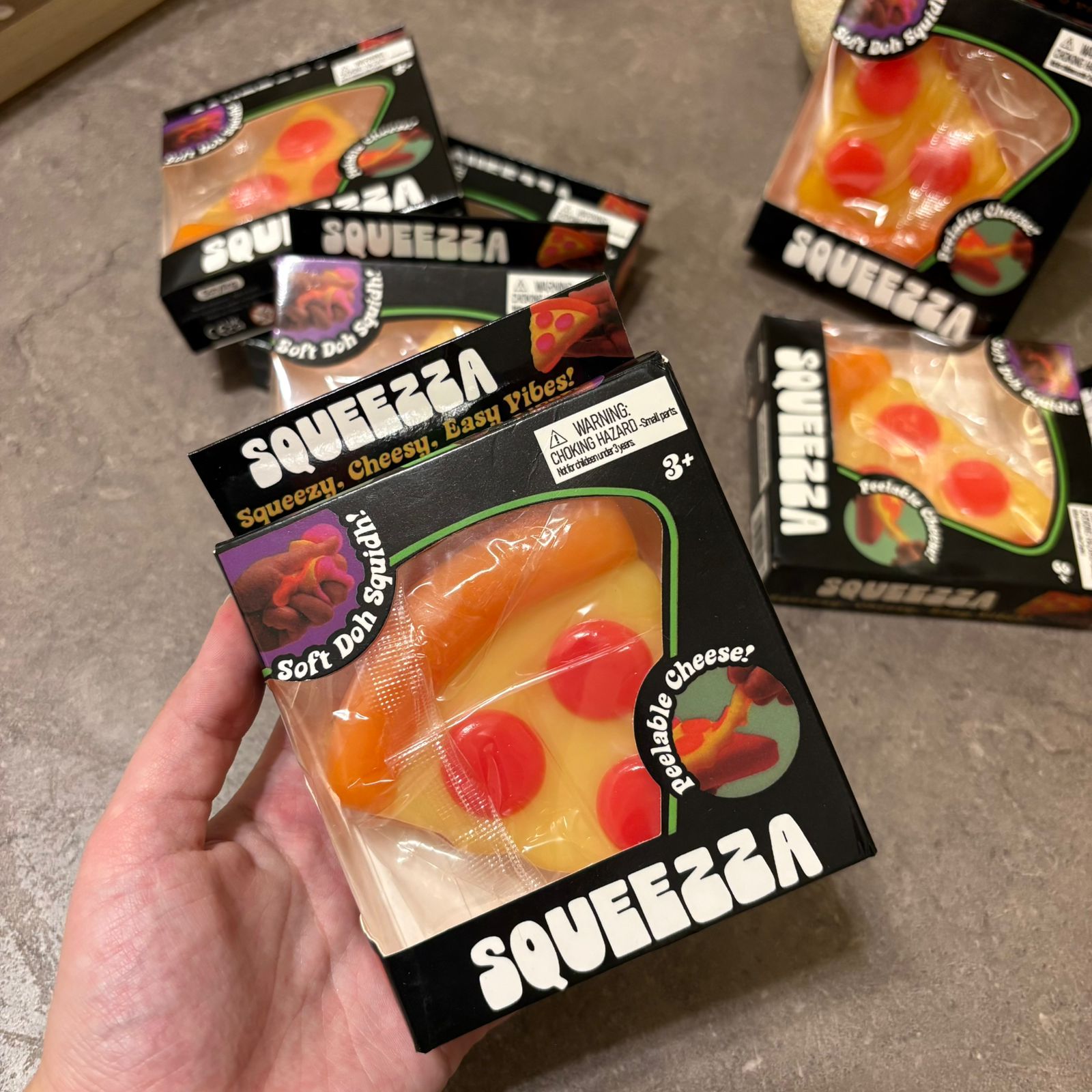 A hand holds a package of Squeezza Soft Dough Squeezies in the shape of a cheese pizza slice, with several other packages scattered on a gray floor in the background.