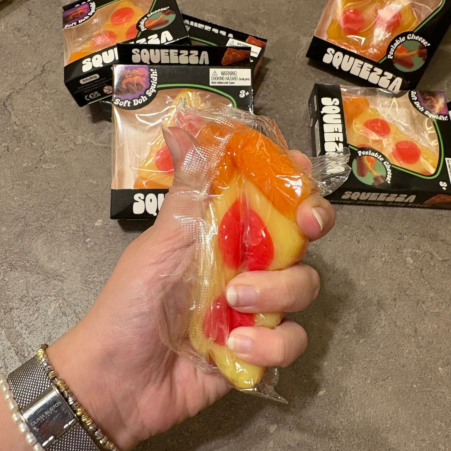 A hand holding a packaged sour peach squishy toy, with several boxed sour peach squishies on a gray concrete floor in the background.