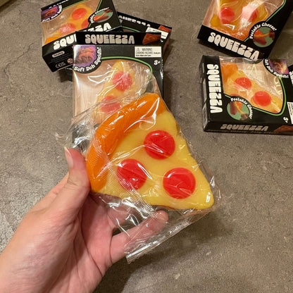 A hand holds a packaged slice of pizza-shaped squishy toy in front of several similar packaged toys on a gray concrete floor. The packaging reads SQUEEZZA.