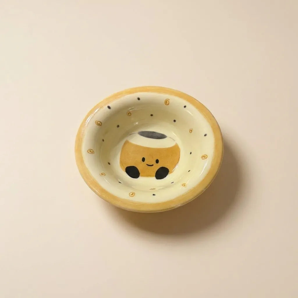 A small hand-painted ceramic bowl with a cute coffee cup character printed inside, set against a plain beige background.