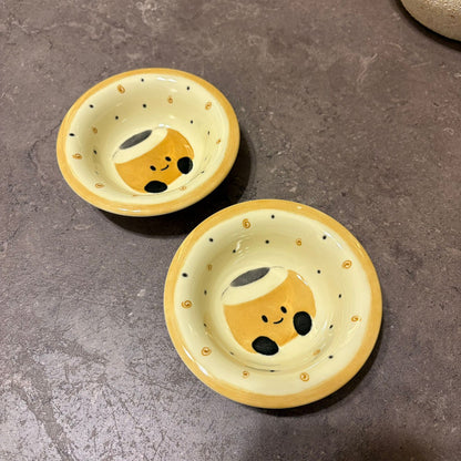 Two small round ceramic bowls with a yellow and cream color scheme, decorated with a cute cartoon face and gold dots, set on a dark gray stone surface.