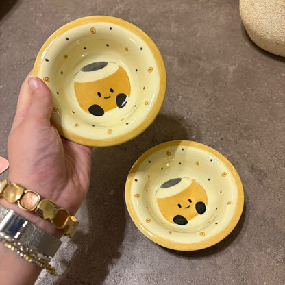 A hand holds a small yellow ceramic bowl with a cute cartoon character printed inside, with a second matching bowl resting on a gray stone surface below.