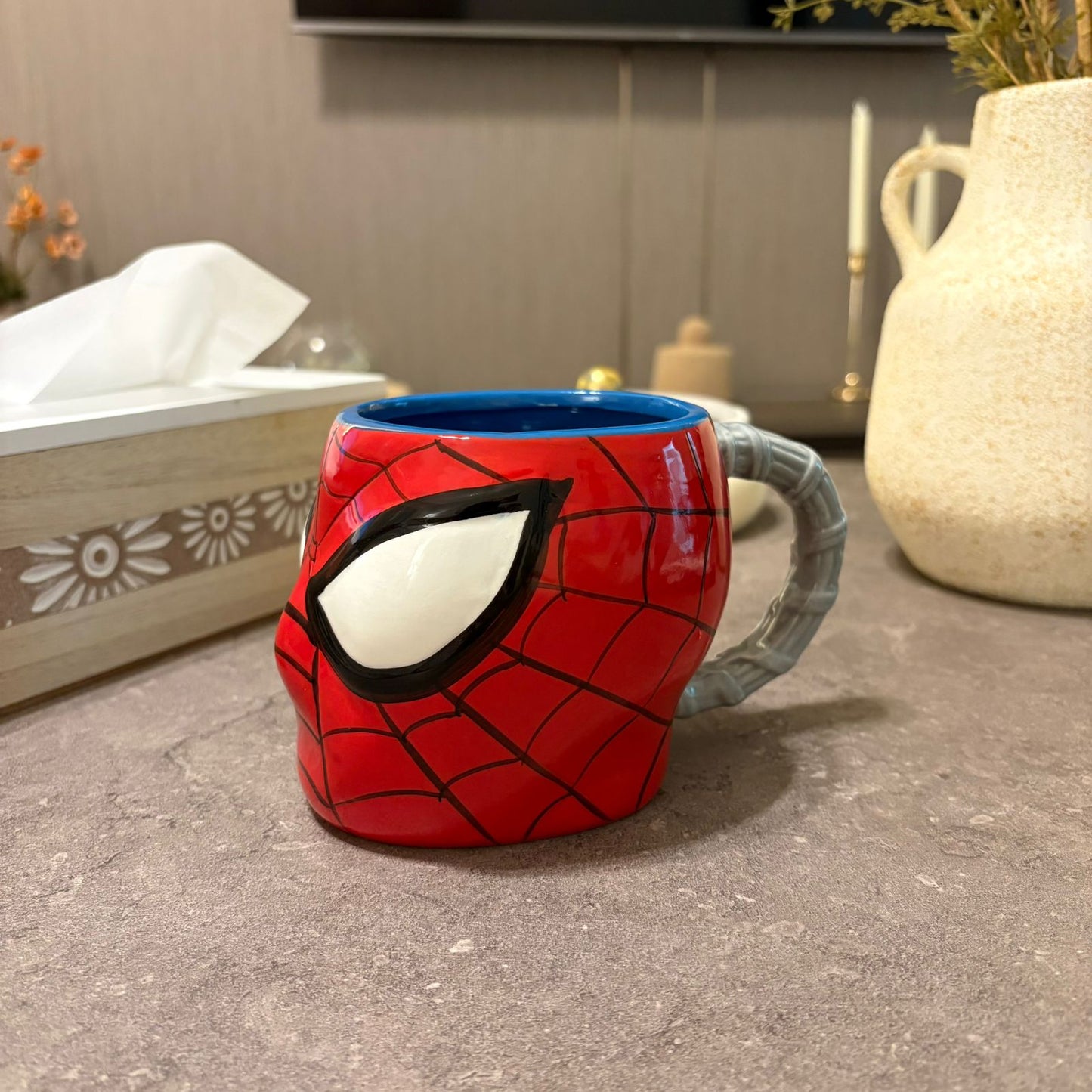 A red and black Spider-Man-themed ceramic mug with a gray webbed handle, placed on a gray countertop in a kitchen setting.