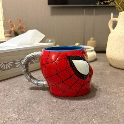 A red and black Spider-Man themed ceramic mug with a silver handle, placed on a gray countertop in a kitchen setting.