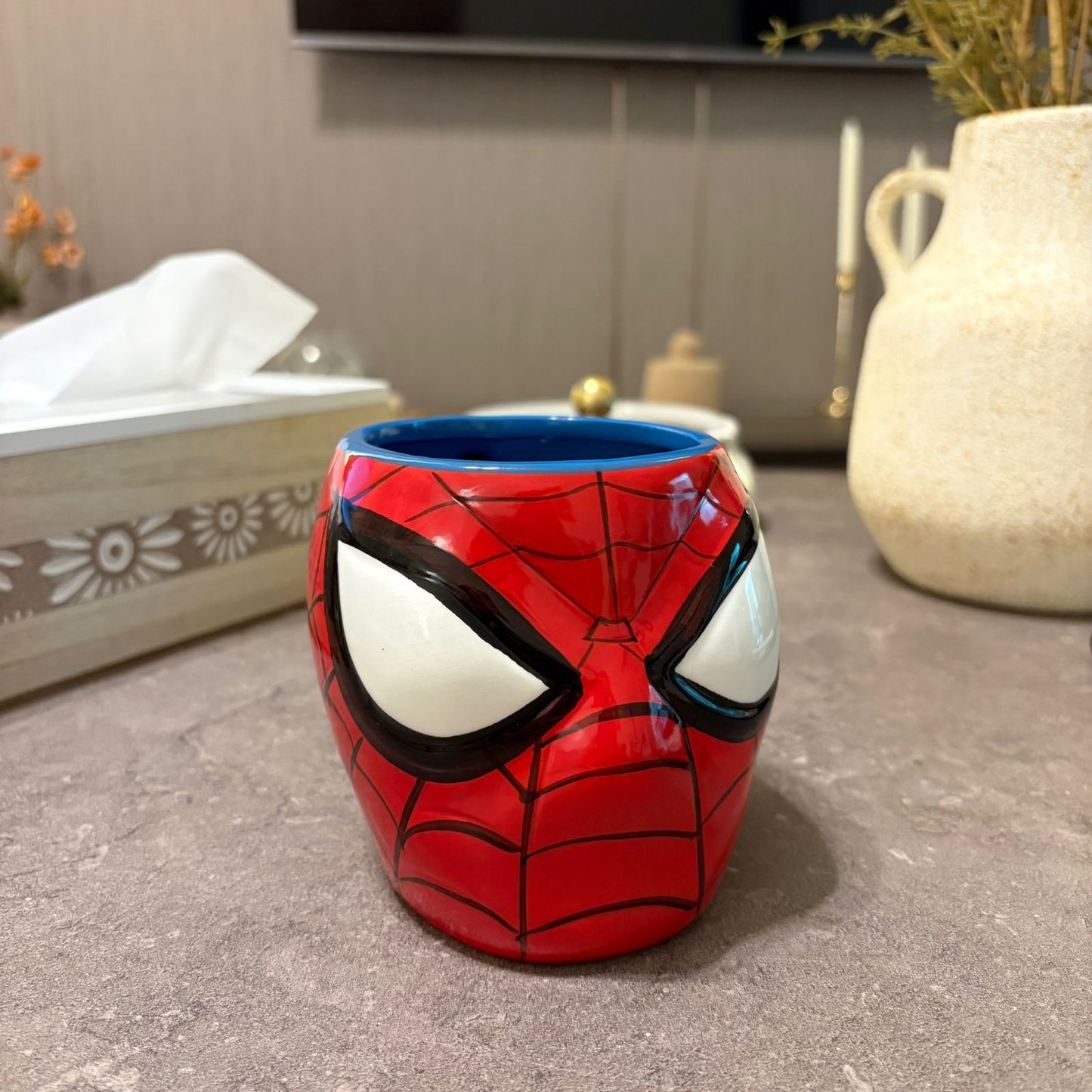 A ceramic mug shaped like Spider-Man's face, featuring red and black web patterns, large white eyes, and a blue interior, set on a gray countertop with a tissue box and vase in the background.