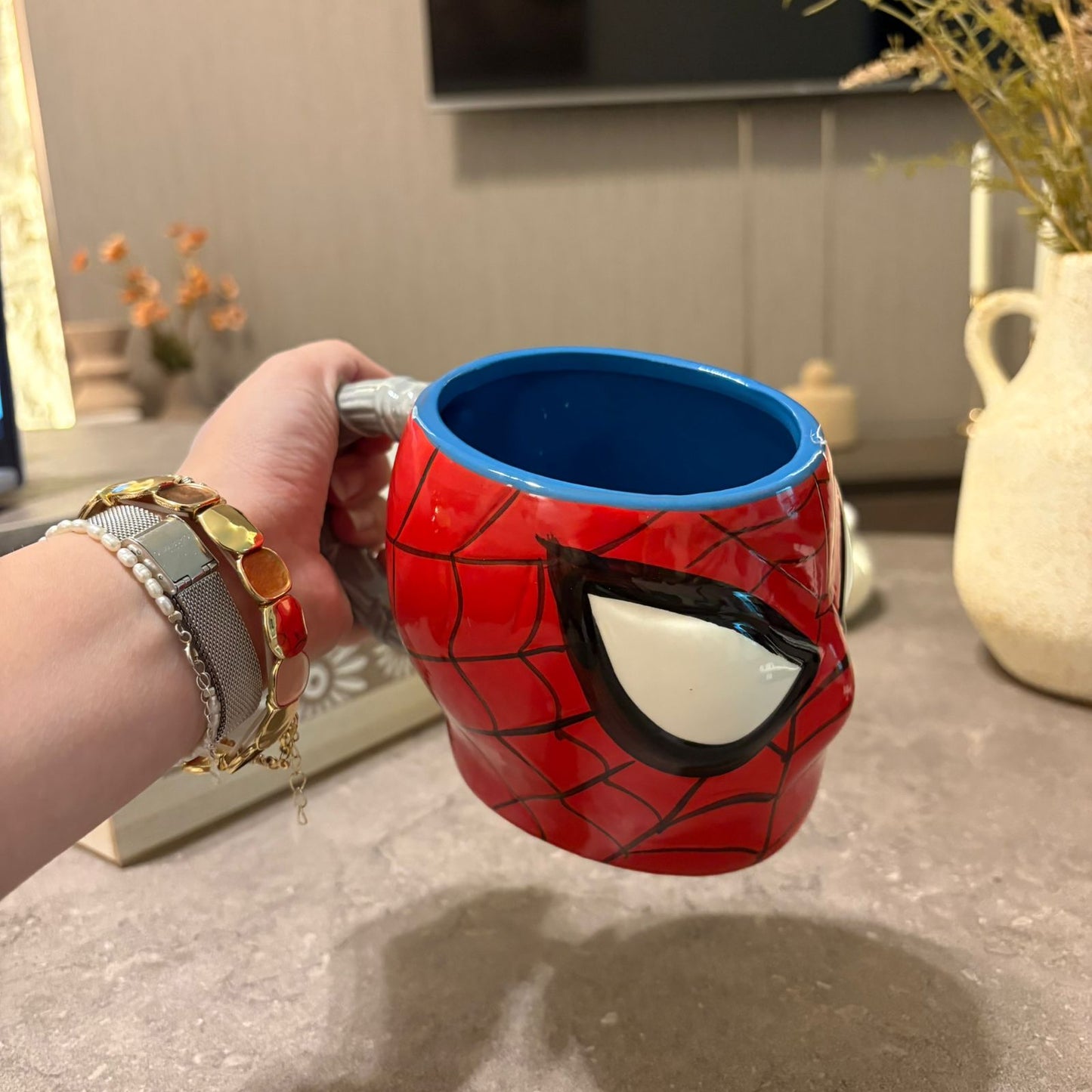 A hand holding a red and blue Spider-Man themed ceramic mug with a stylized eye design on the side, set against a blurred indoor background.
