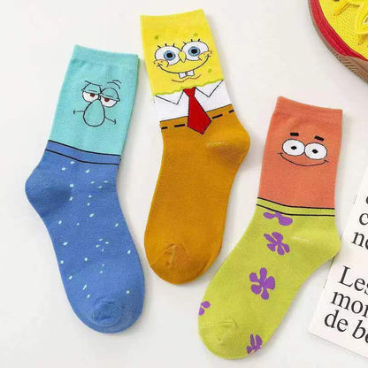 Three cartoon-print ankle socks laid flat on a white surface: a light blue sock with a green character on the cuff and blue foot, a yellow sock with a SpongeBob-inspired face and red tie on the cuff and orange body, and a peach-orange sock with a face on the cuff and purple flowers on the foot.