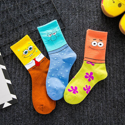 Three colorful cartoon socks laid flat on a dark textured surface. One sock is yellow and orange with SpongeBob-style face details, one is blue and light blue with a sleeping face, and one is lime green and orange with pink flower details.