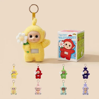 A yellow Teletubbies character plush keychain holding a white flower stands next to its branded box on a warm beige background. Smaller versions of the same keychain in other colors are lined up along the bottom.