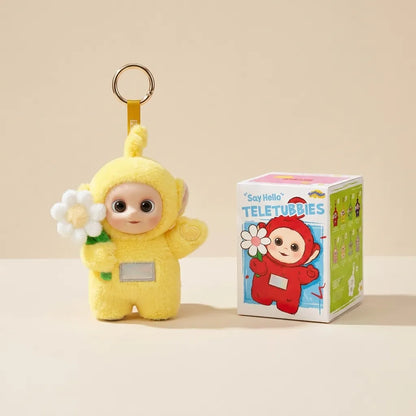 A yellow Teletubbies plush keychain character holding a white flower, shown next to its branded retail box on a warm beige background.