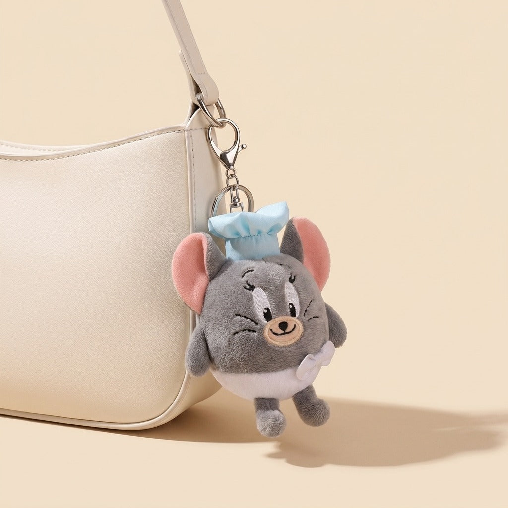 A plush character keychain shaped like a gray mouse with pink ears, a light blue bow, and a white apron, hanging from a cream-colored handbag strap against a warm beige background.