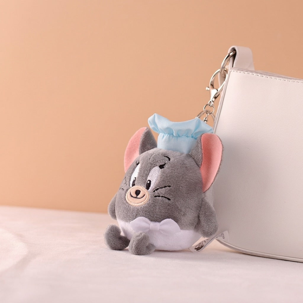 A plush character keychain shaped like a gray cat wearing a light blue hat and white bow, shown resting beside a white handbag on a soft beige surface.
