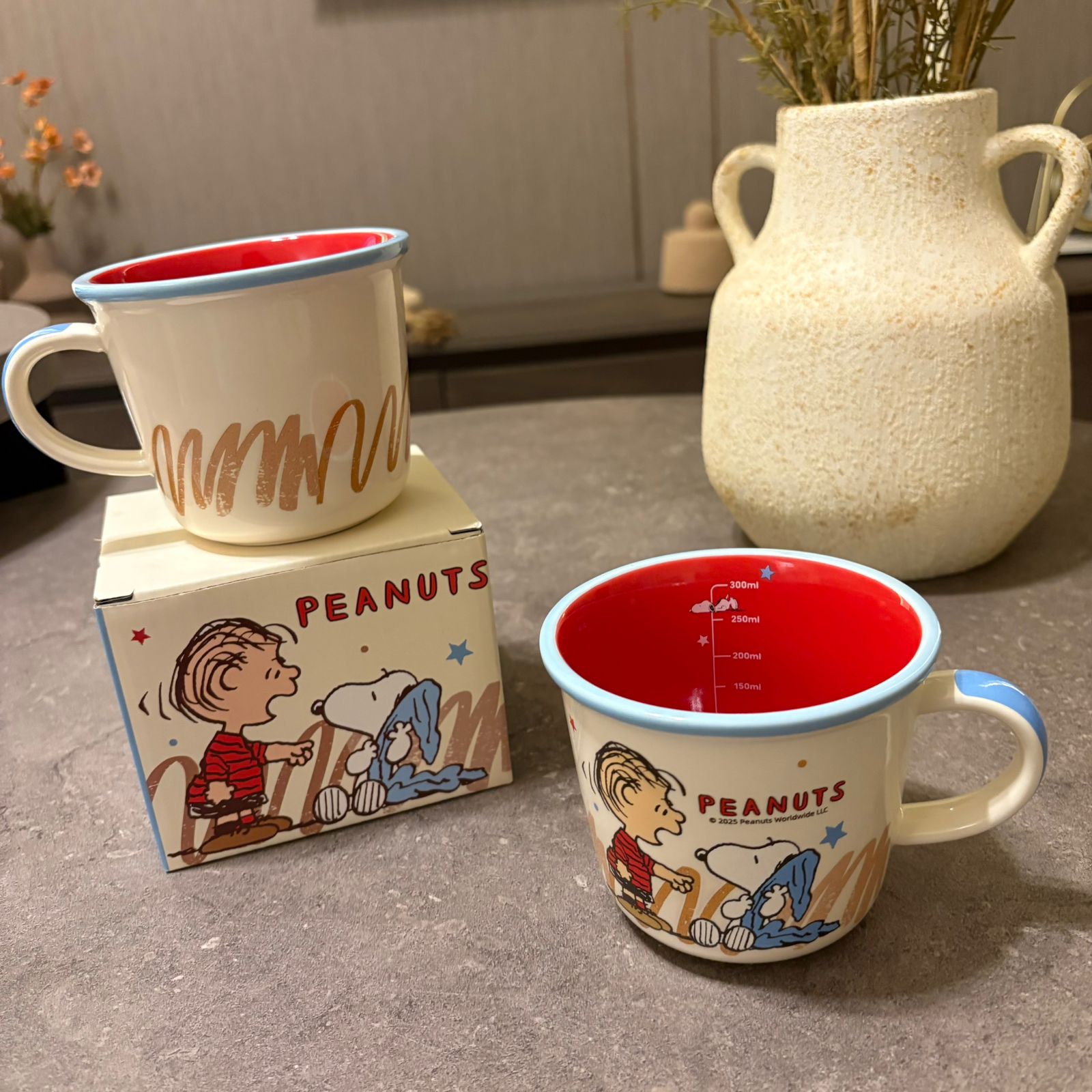 Two Peanuts-themed enamel mugs on a gray tabletop, one standing on its branded box and one displayed beside it, with a ceramic vase in the blurred background.