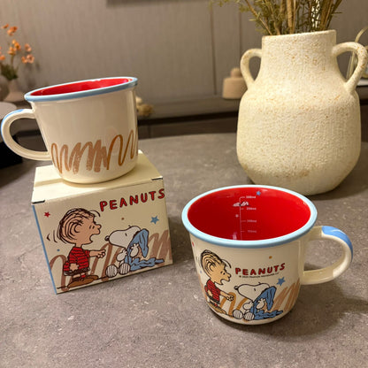 Two Peanuts-themed enamel mugs on a gray tabletop, one standing on its branded box and one displayed beside it, with a ceramic vase in the blurred background.