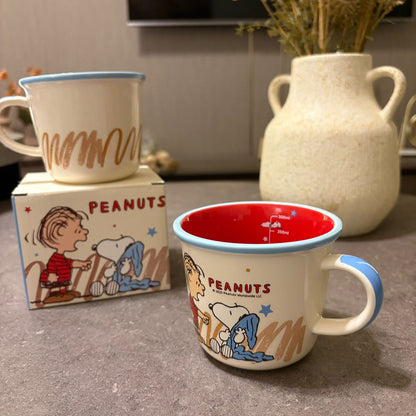 A Peanuts enamel mug with Snoopy and Charlie Brown illustration, blue handle and rim, and a red interior, shown in front of its branded box with another mug in the background. A cream ceramic vase with dried stems sits on a gray tabletop.