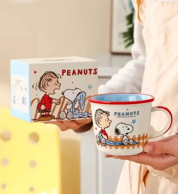 A person holds a white ceramic Peanuts mug with red trim and cartoon characters, with the matching Peanuts box behind it in a softly lit indoor setting.