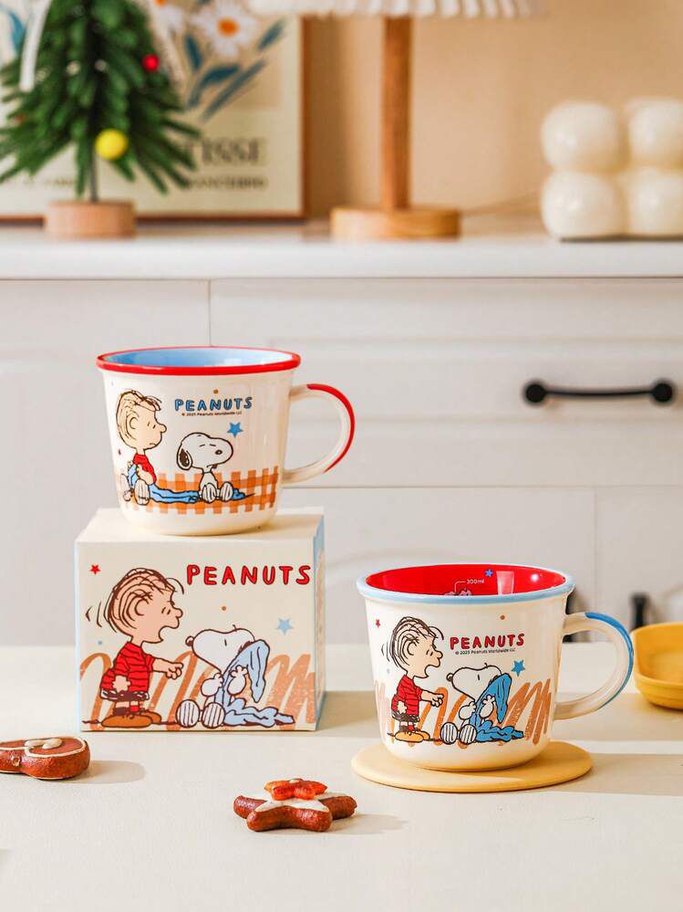 Two Peanuts enamel mugs with cartoon character illustrations are displayed on a kitchen counter, one resting on its box and the other on a coaster. The mugs have red rims and handles, with one mug featuring a blue handle.