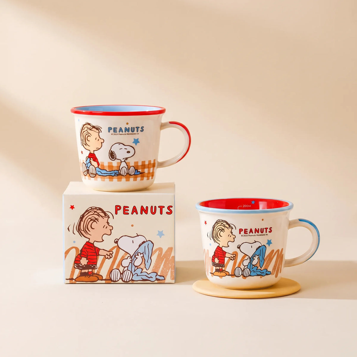 Two white Peanuts mugs with red and blue trim, featuring cartoon illustrations of Charlie Brown and Snoopy, shown with their branded box on a warm beige background.