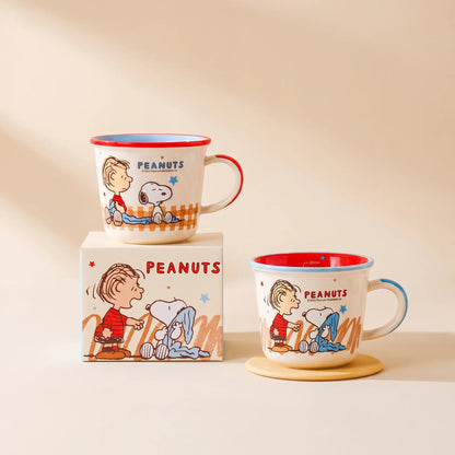 Two white Peanuts mugs with red and blue trim, featuring cartoon illustrations of Charlie Brown and Snoopy, shown with their branded box on a warm beige background.