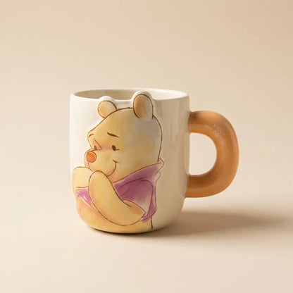 A ceramic mug featuring Winnie the Pooh on the front and a tan handle, photographed on a plain beige background.