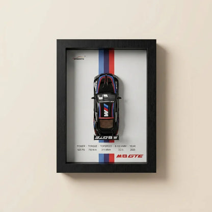 A framed automotive poster featuring a top-down view of a black BMW M2 GTE race car with red, white, and blue racing stripes, mounted on a light beige wall.