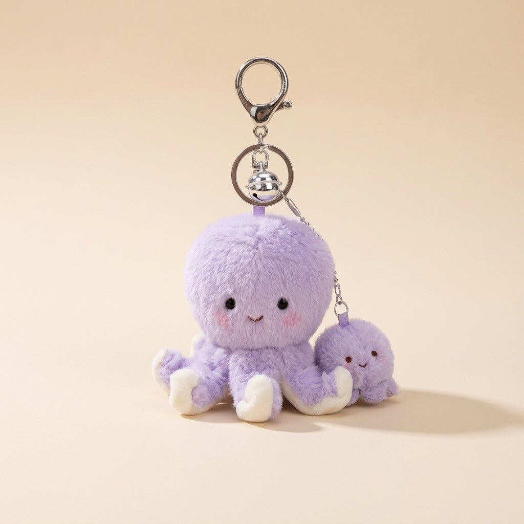A cute lavender plush octopus keychain with a small matching plush charm, attached to a silver metal clasp and bell, set against a solid beige background.