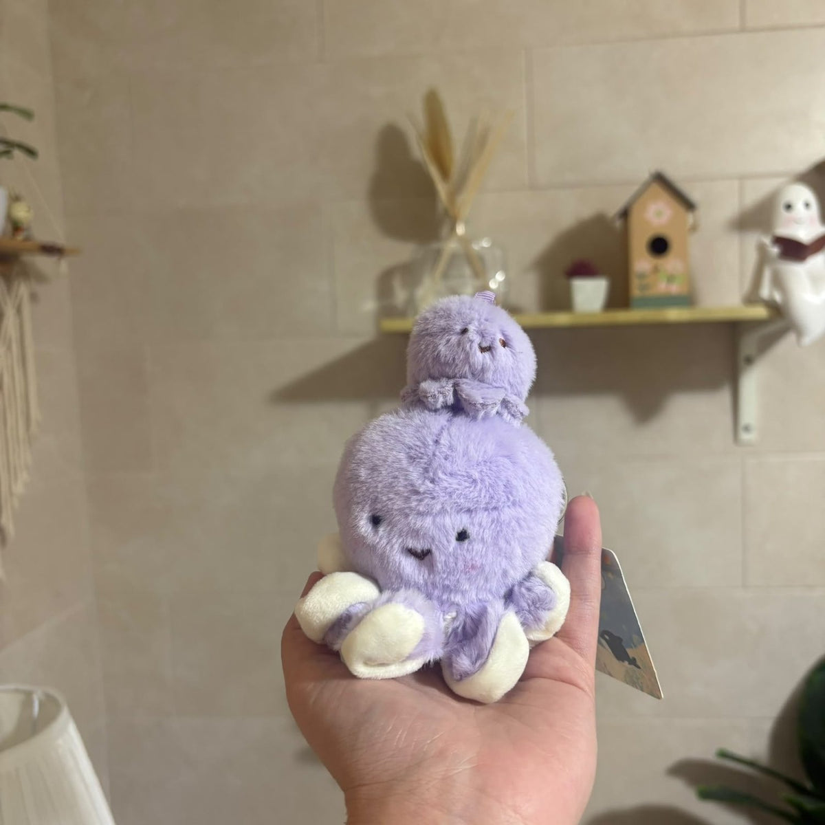 A small lavender plush octopus toy with cream-colored feet is held in a person's hand, with a beige tiled wall and shelf decorations in the background.