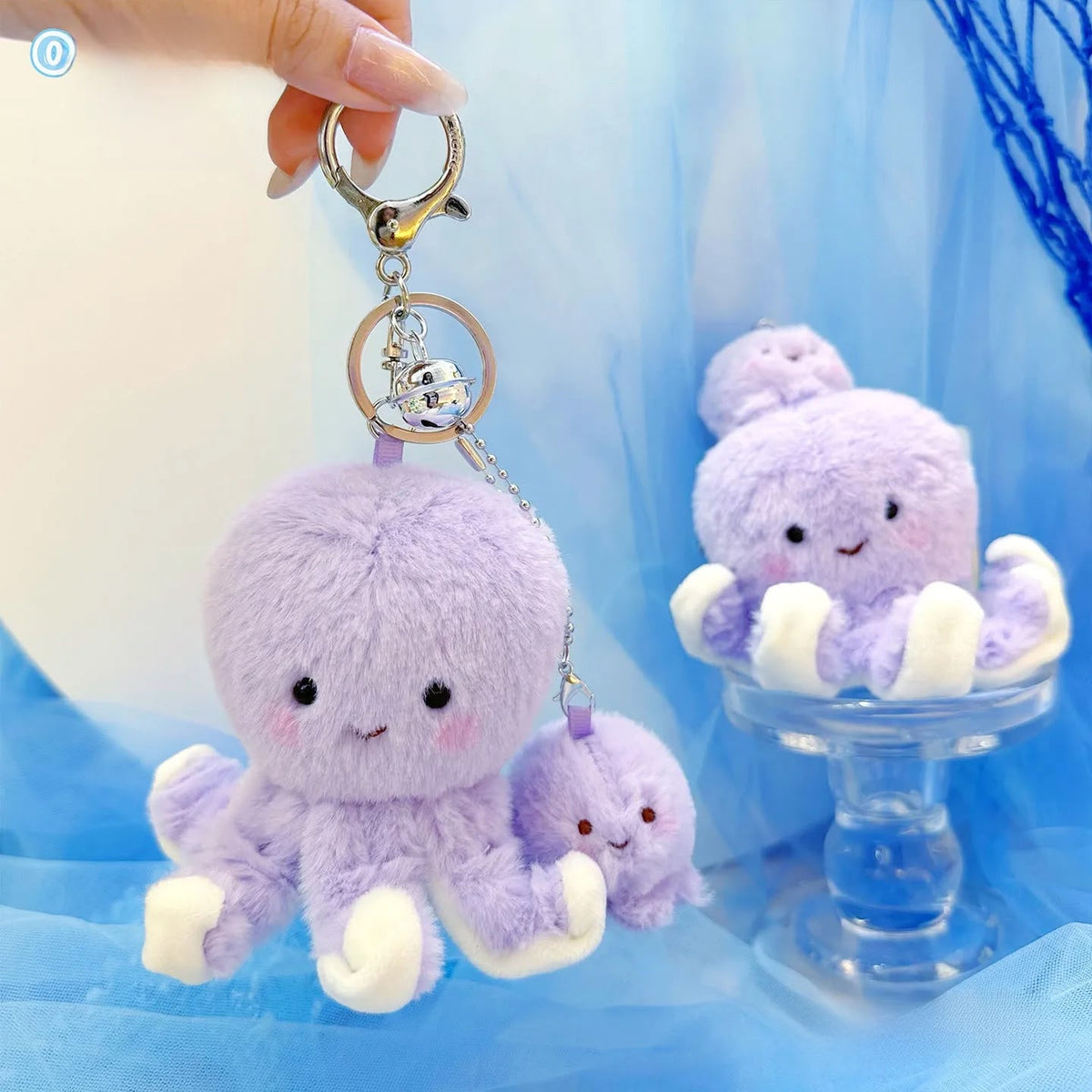 A lavender plush octopus keychain with a small matching plush charm, held by a hand against a soft blue tulle backdrop. The keychain has a gold-tone split ring and chain, and the octopus has white feet and pink cheeks.
