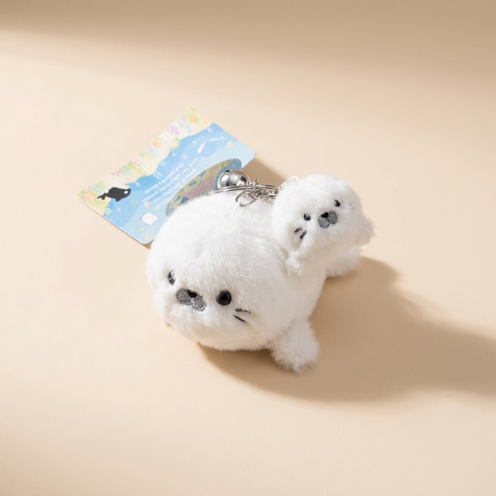 A plush white seal keychain charm with a small attached card, photographed on a warm beige studio background.