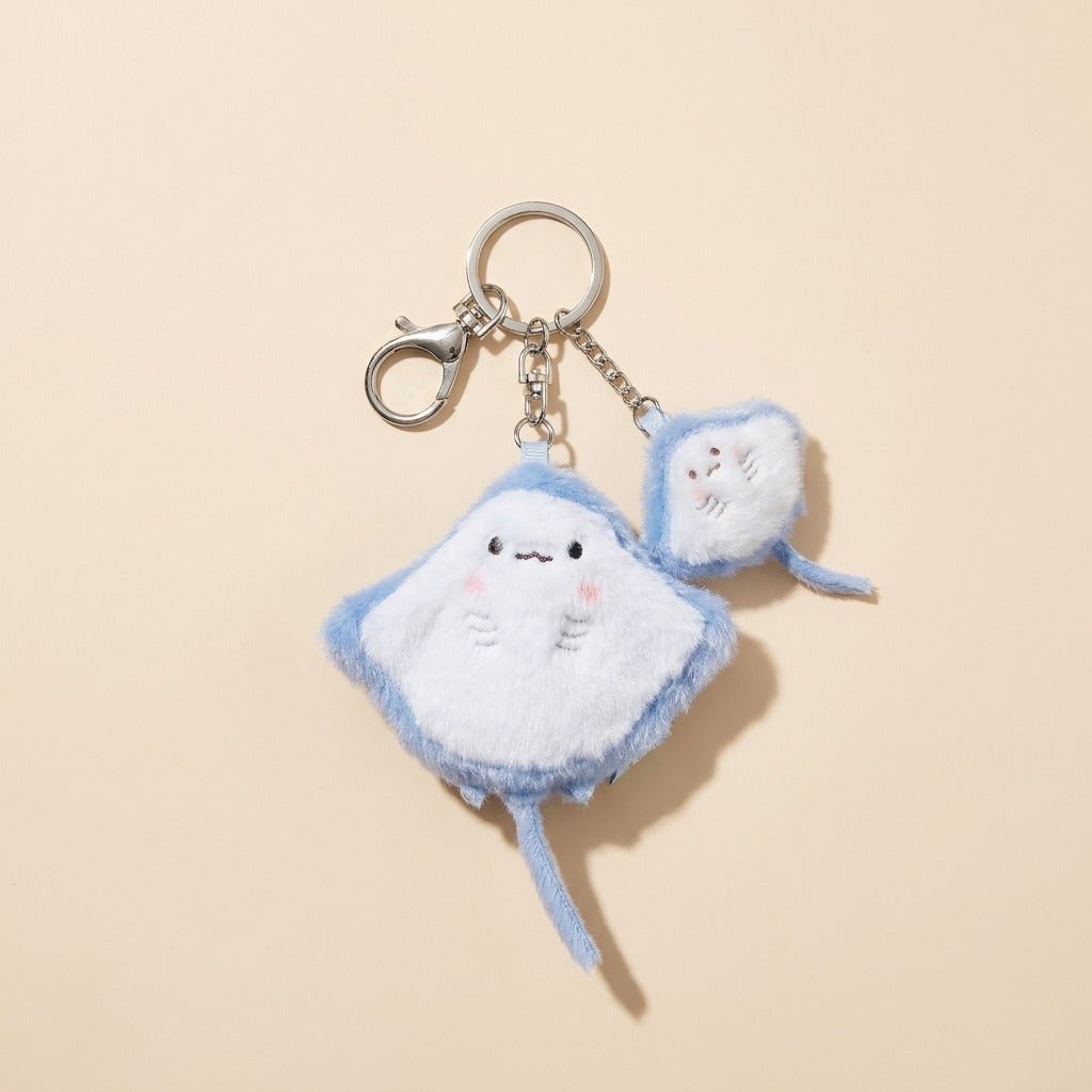 A cute plush keychain featuring a light blue and white stingray character with a small matching pouch, attached to a silver metal clasp on a warm beige background.
