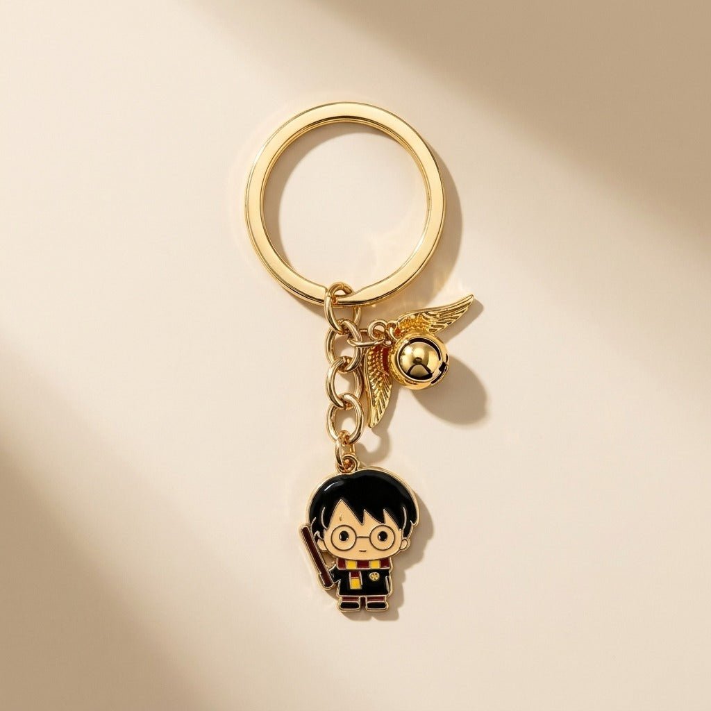 A gold-tone metal keychain featuring a small character charm with black hair, round glasses, and a wand, alongside a gold winged bell charm, photographed on a warm cream background.