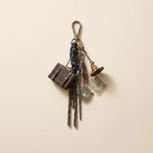 A vintage-style keychain charm set featuring miniature accessories including a brown briefcase, a small bell, a compass, a ticket, and patterned keys, hanging from a silver carabiner on a warm beige background.