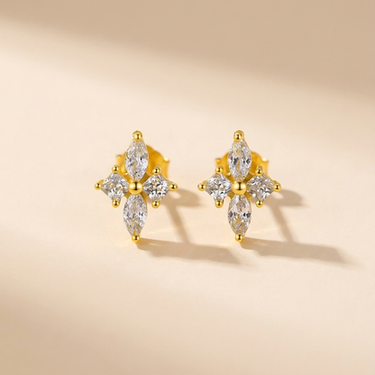 A pair of gold stud earrings with marquise-cut clear gemstones arranged in a floral or cross-like cluster, photographed on a warm beige background with soft shadows.