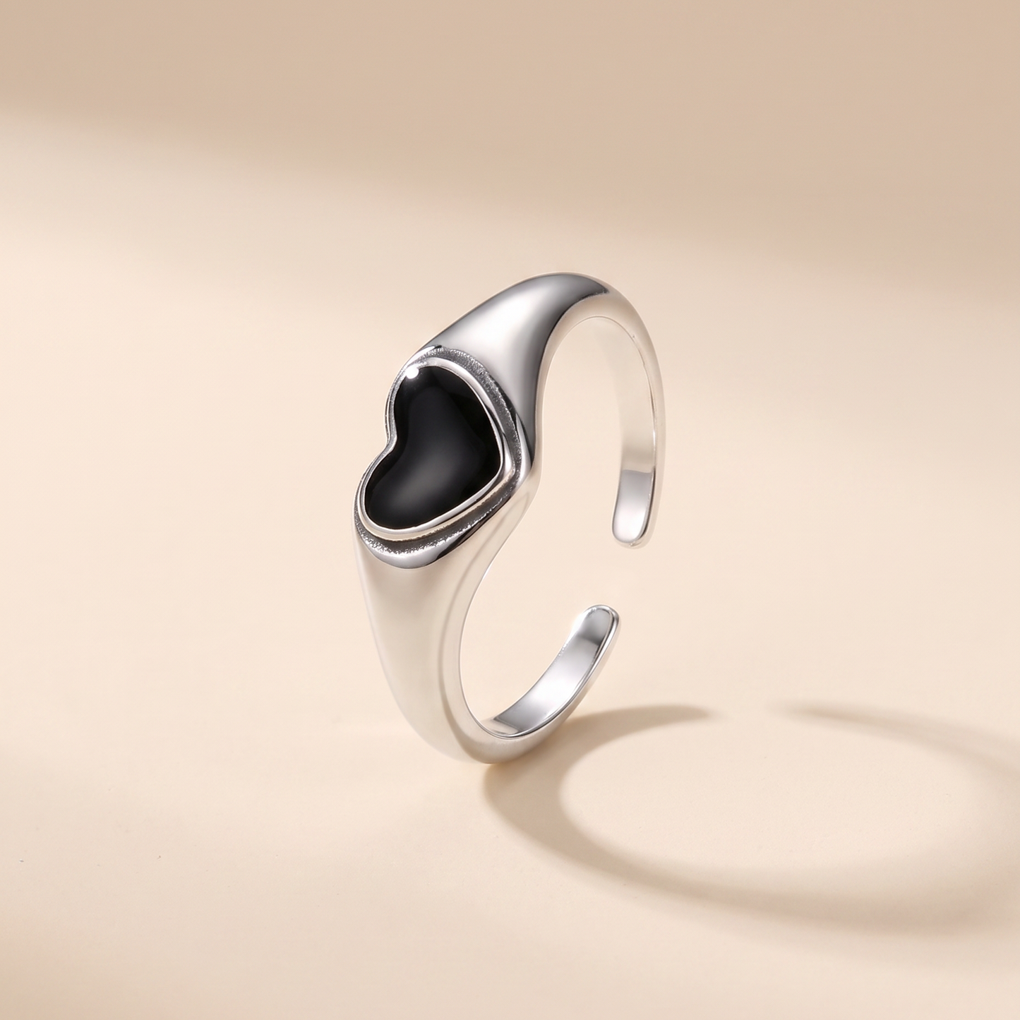 A polished silver-toned ring with a heart-shaped black inlay, shown on a warm beige background with a soft shadow.
