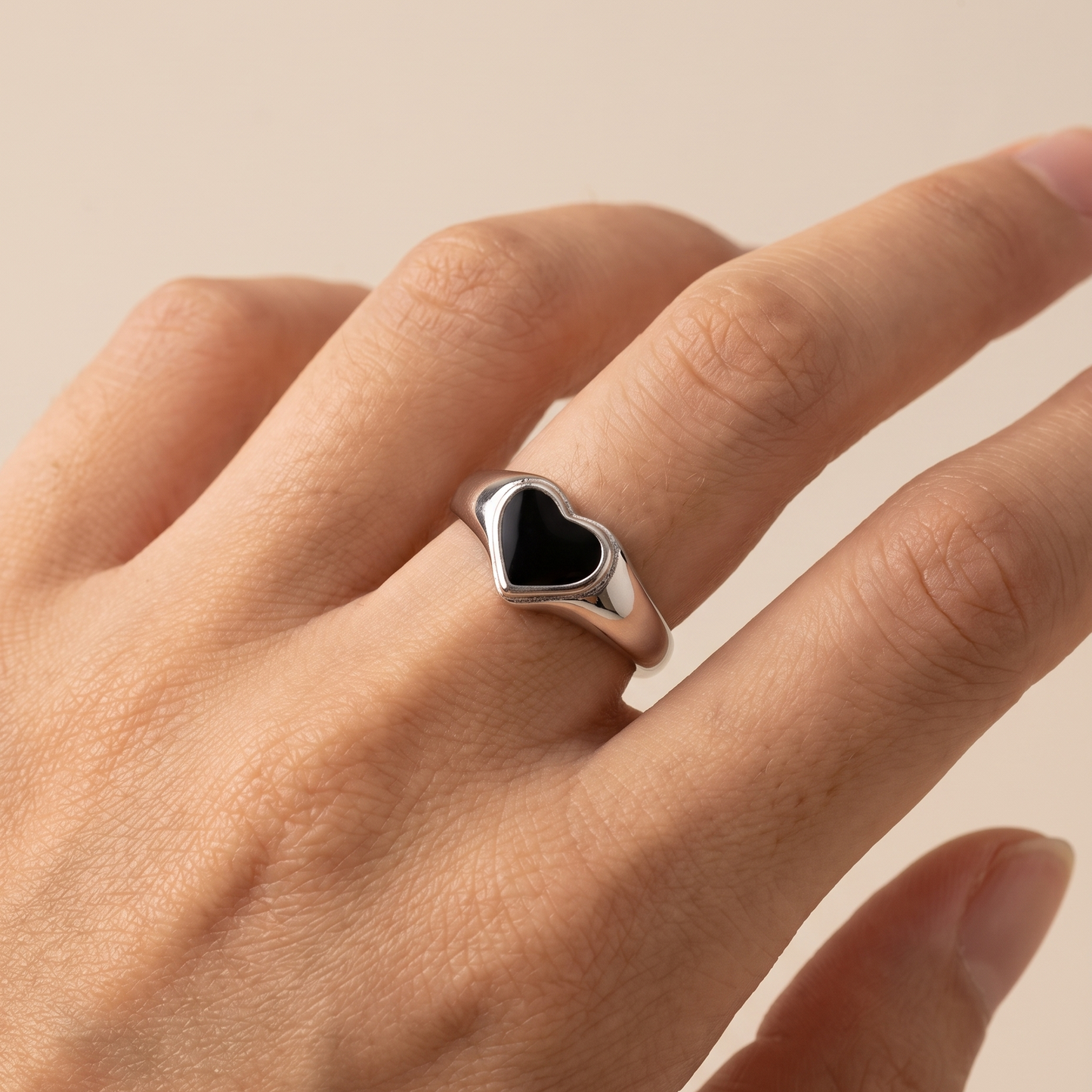 A silver ring featuring a polished heart-shaped black inlay, shown worn on a person's finger against a soft beige background.