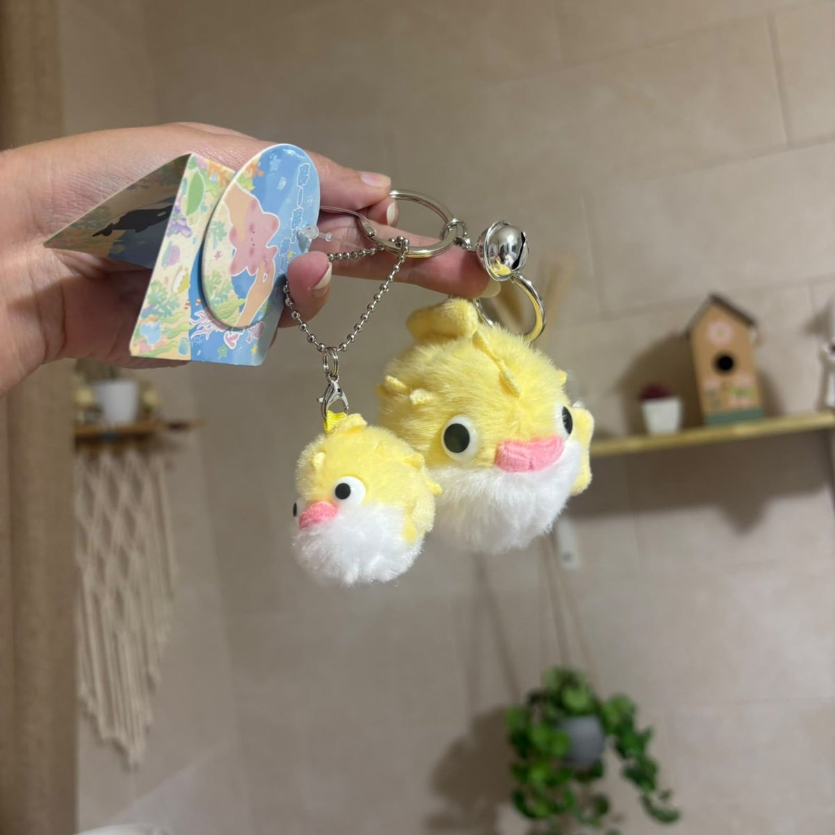 A hand holds two small yellow plush duck keychains with white fluffy trim and pink beaks, attached to silver chains with a bell, in front of a softly blurred indoor room background.