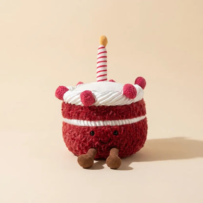 A plush cake toy with red fuzzy frosting, white frosting, a striped candle, and small brown legs, photographed on a warm beige studio background.