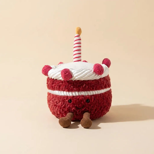 A plush cake toy with red fuzzy frosting, white frosting, a striped candle, and small brown legs, photographed on a warm beige studio background.