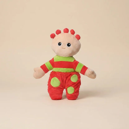 A small plush toy character standing against a plain warm beige studio background. The character has a cream face and hands, red hair spikes, a red-and-green striped top, and red pants with green circular patches.